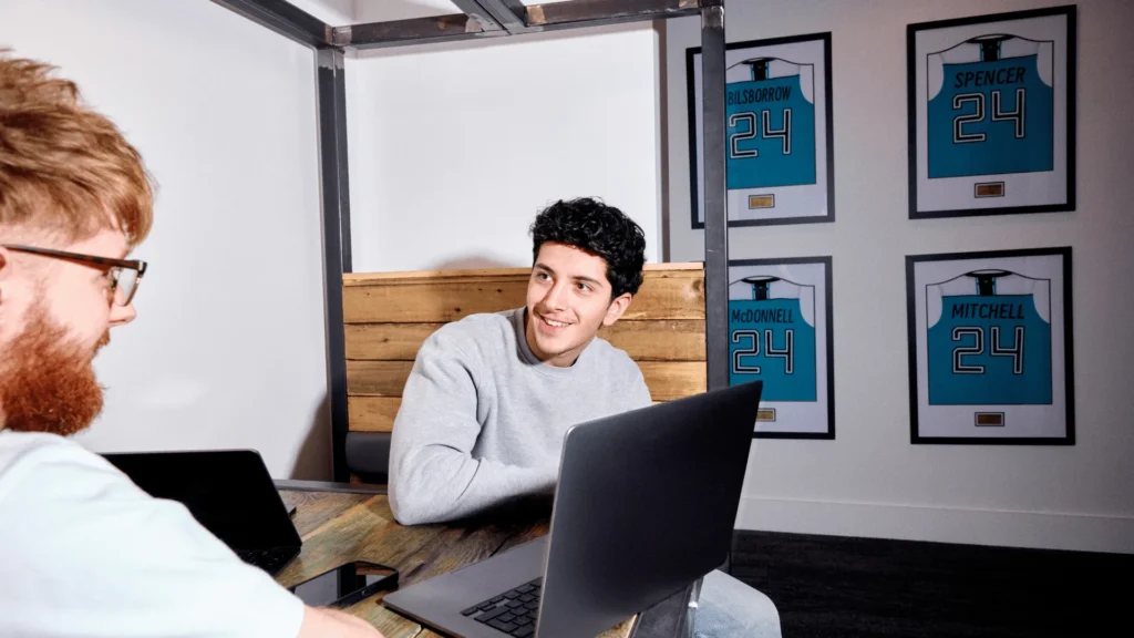 Two young male professionals in a modern office, one smiling at the other who is sitting with a laptop, representing Australian business owners discussing how to lodge a Business Activity Statement (BAS).