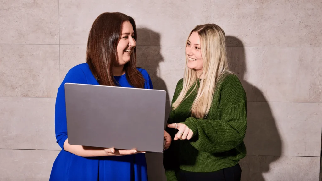 Two female colleagues smiling and looking at a laptop together, representing a positive manager and employee conversation about career growth and promotion criteria.