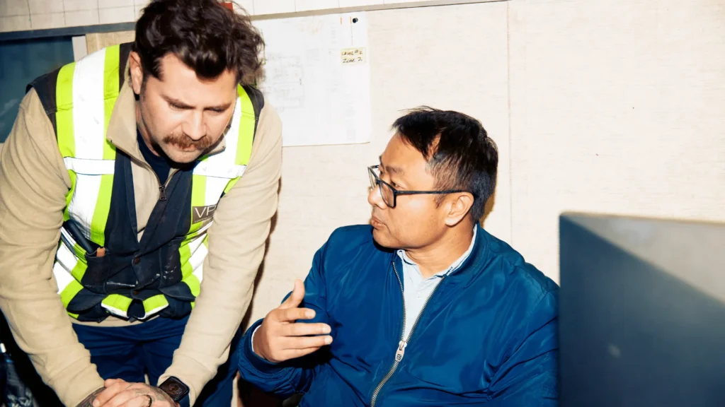 A construction worker in a high-visibility vest assisting a colleague at a computer desk, demonstrating how an onboarding buddy program supports training and integration for deskless and site-based employees.