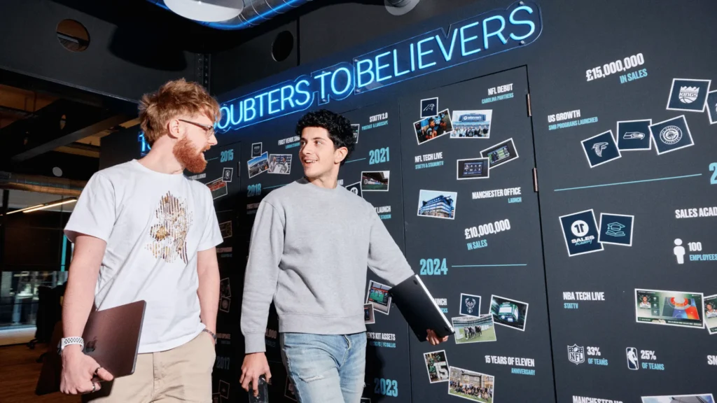 Two young professionals walking and talking in a modern, branded office hallway, illustrating an onboarding buddy giving a new hire an informal office tour and introducing them to the company culture.