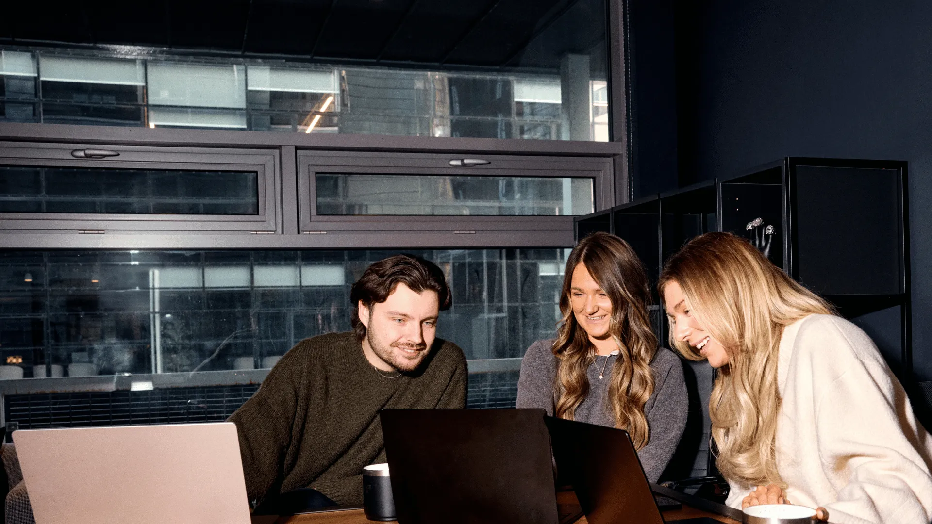 Three smiling colleagues collaborating around laptops in a modern office, representing how a successful onboarding buddy program helps new hires quickly integrate and build relationships with their team.