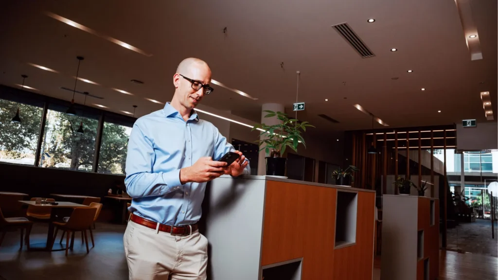 A smiling male professional wearing glasses and a blue button-down shirt checking his smartphone in a modern office space, representing an Australian SME owner reading updates on the 2026–27 federal budget.