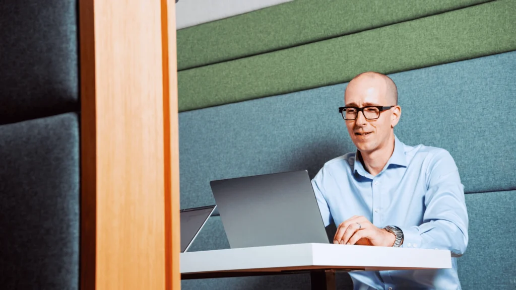 A man in a light blue shirt sits at a desk working on a laptop. The environment is modern with muted blue and green wall panels, evoking a calm, focused ambiance.