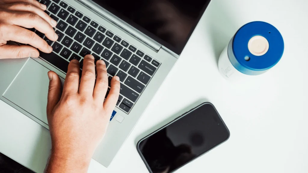 A top-down view shows a person typing on a laptop on a white desk, next to a smartphone and a blue-and-white cup.