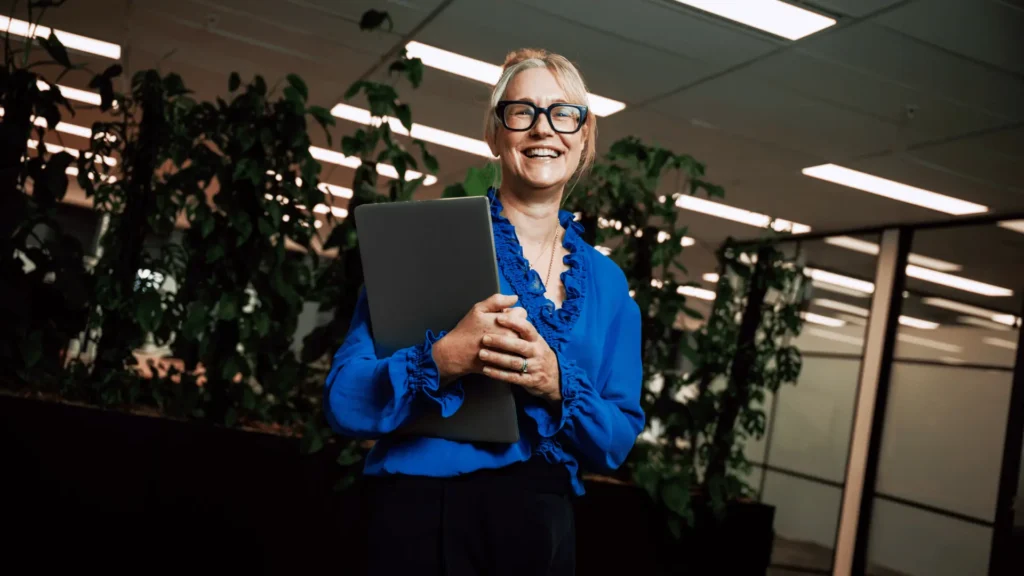 A woman in a bright blue blouse holds a laptop and smiles warmly in an office with indoor plants. The scene conveys a positive and professional atmosphere.