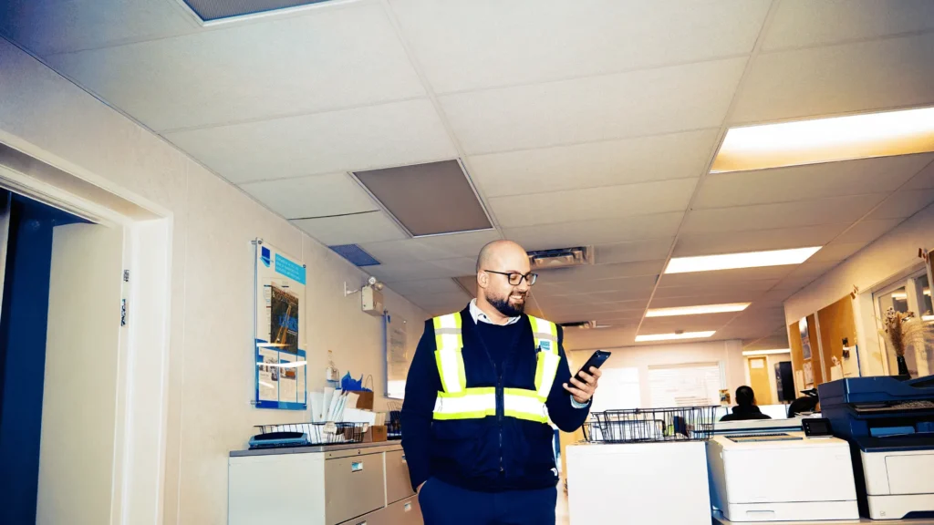A smiling male contractor wearing a high-vis vest and safety glasses checking his smartphone in an office hallway, representing subcontractors receiving their superannuation payments.