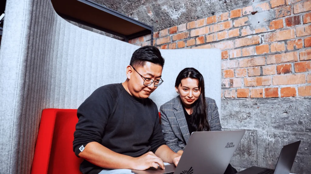 A man and a woman sitting together on a red office couch, collaboratively reviewing employment agreements on a laptop to master their contract management process.