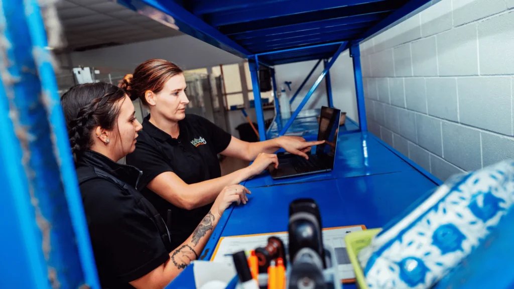 Two female warehouse employees collaborating over a laptop on a blue industrial shelf, illustrating how employers manage and review superannuation payments for their contractors.