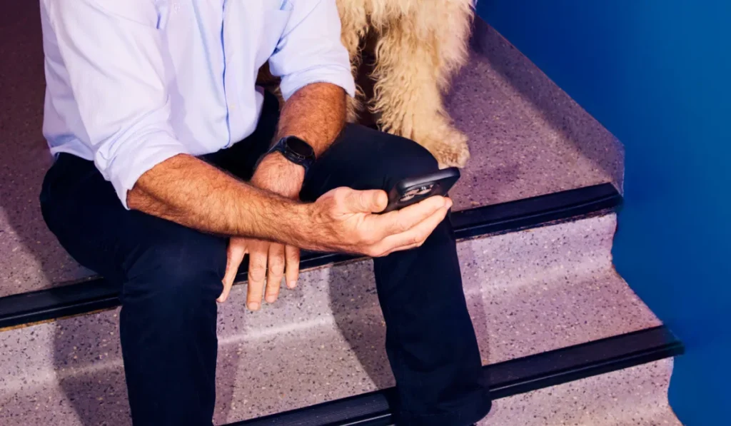 A cropped image showing a person sitting on speckled gray stairs with a solid blue wall in the background. The person is wearing a light blue shirt and dark trousers, holding a black smartphone in their hands. The fluffy fur of a light-colored dog is visible sitting on the step right next to them.