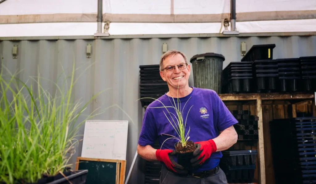 A smiling man wearing glasses, a purple Kaipātiki Project t-shirt, and red gardening gloves holds a small potted plant in a greenhouse setting. Behind him are stacks of black plastic planting trays, a green bin, and a corrugated grey wall.