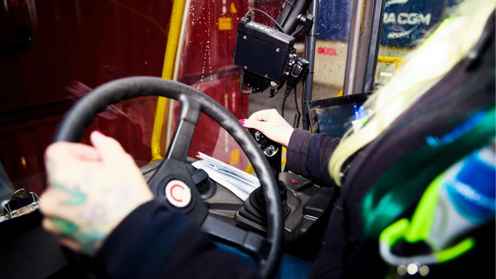 Close-up view of a driver's hands with tattoos operating the steering wheel and joystick control lever inside a heavy machinery cabin.