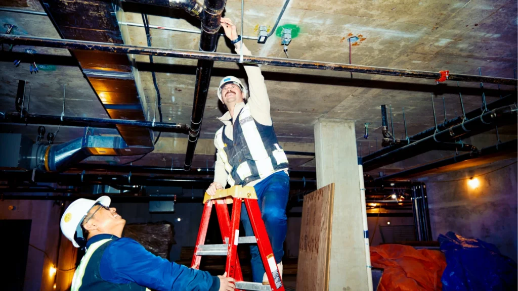 Two workers wearing hard hats inspect overhead piping in a basement, with one man standing on a red stepladder pointing at a fixture.