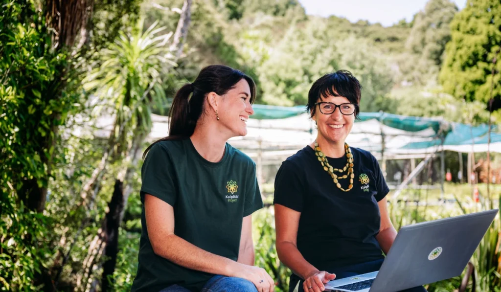 Two smiling women sitting outdoors at a rustic wooden table on a sunny day. The woman on the right is using a silver laptop. Behind them is a grassy area with plants, trees, and a blackboard displaying a handwritten welcoming message.