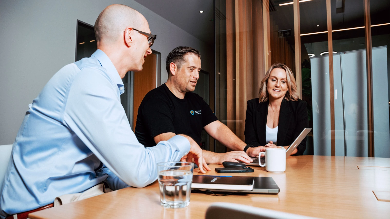 Three professionals gather around a wooden conference table, looking together at a laptop screen during a collaborative workplace meeting.