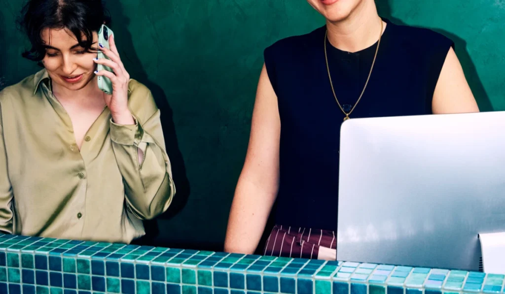 Two women standing behind a reception desk covered in teal square tiles against a dark green wall. The woman on the left is wearing a silky olive-green shirt and talking on her smartphone, while the woman on the right wears a black sleeveless top and stands next to a silver computer monitor.