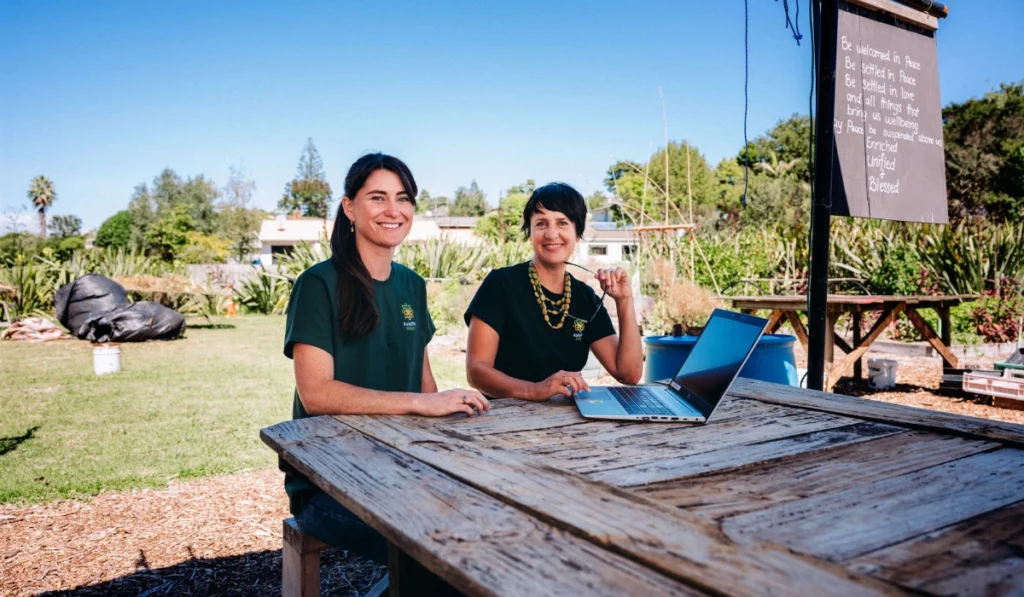 Two smiling women sitting outdoors at a rustic wooden table on a sunny day. The woman on the right is using a silver laptop. Behind them is a grassy area with plants, trees, and a blackboard displaying a handwritten welcoming message.