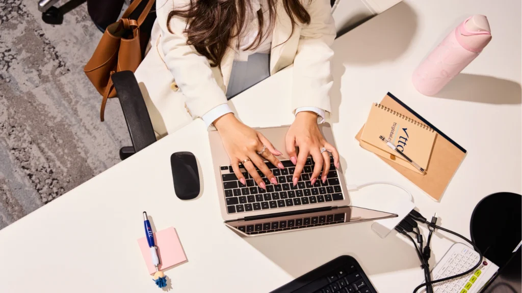 A top-down view of a woman wearing a white blazer typing on a rose-gold laptop on a white desk. The desk is organized with a black mouse, a pink water bottle, notebooks, a pen, pink sticky notes, and various computer cables and accessories.