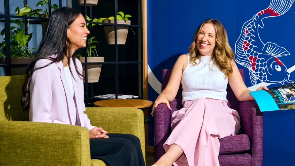 Two women laughing and talking while sitting in colorful armchairs in a modern office space. The woman on the left wears a light purple blazer in a green chair, and the woman on the right wears a white top and pink trousers in a purple chair, holding a colorful brochure. The background features a dark shelf with potted plants and a blue wall with a large white and red koi fish mural.