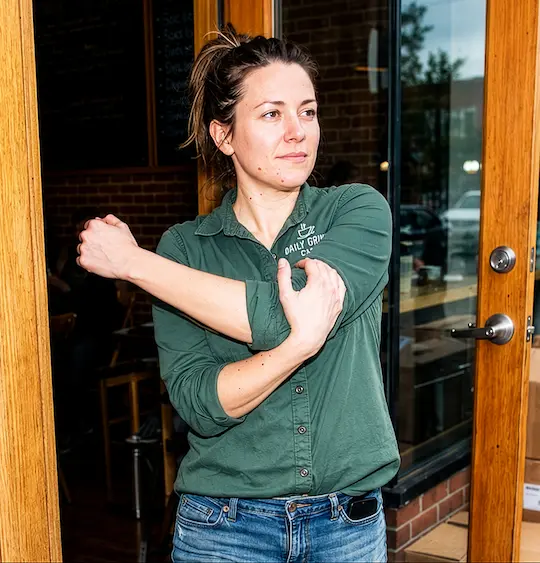 A woman in a green collared shirt stretches her arm while standing in the doorway of a café.