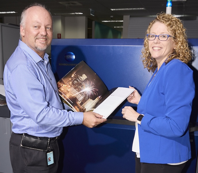 Two smiling colleagues hold open a printed photo booklet in front of a large blue printing machine.