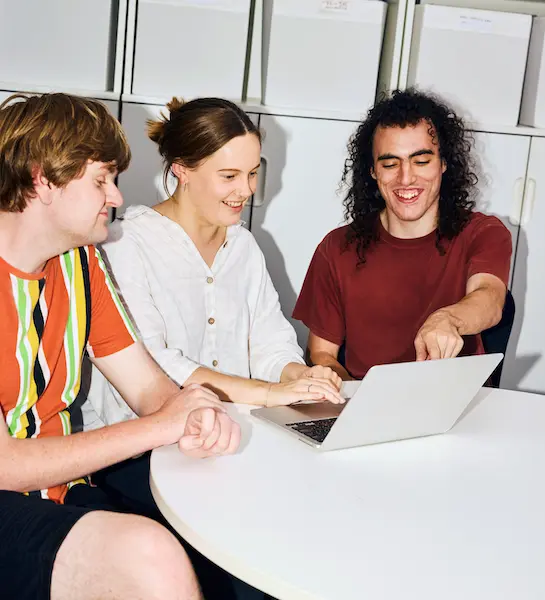 Three smiling colleagues collaborate around a laptop at a desk, with one pointing at the screen.