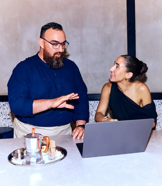 A man in an apron gestures while discussing work on a laptop with a woman at a counter.