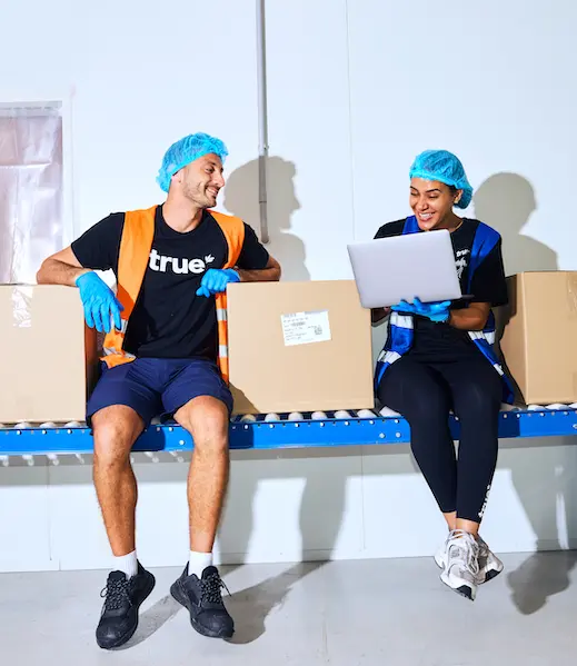 Two smiling warehouse workers in safety gear sit on a conveyor belt next to boxes and look at a laptop together.