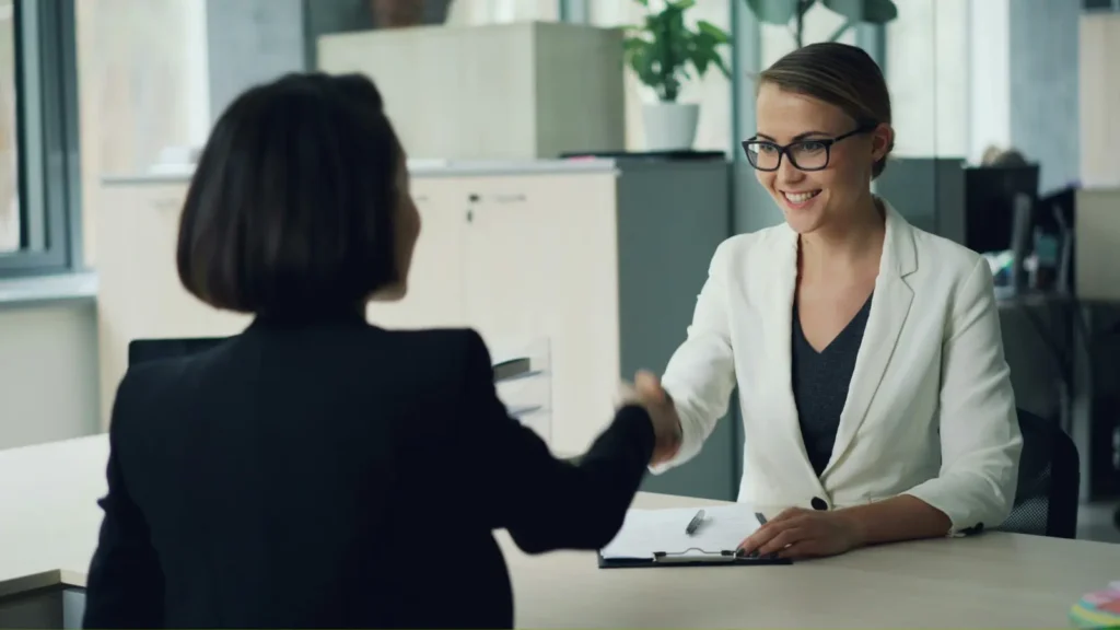 Businesswomen shake hands across a desk in a bright office, with documents nearby; the smiling meeting conveys professionalism and trust.
