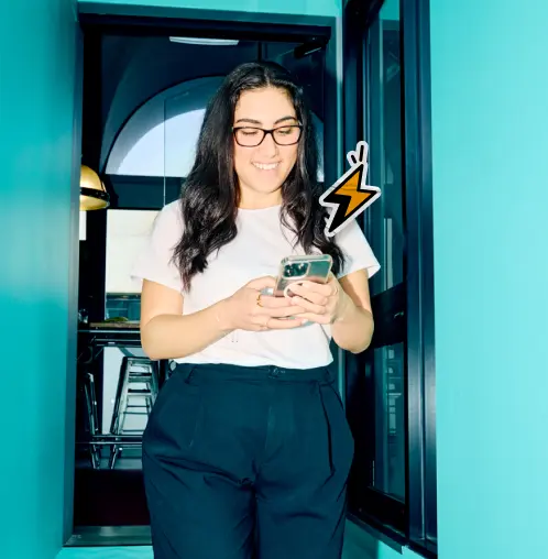 A woman in a bright blue blouse holds a laptop and smiles warmly in an office with indoor plants.