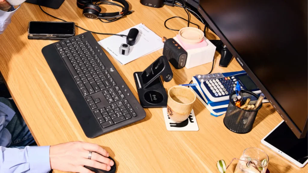 An overhead view of a wooden desk with a person using a computer mouse, a black keyboard, a notebook with a pen, and a cup of coffee.