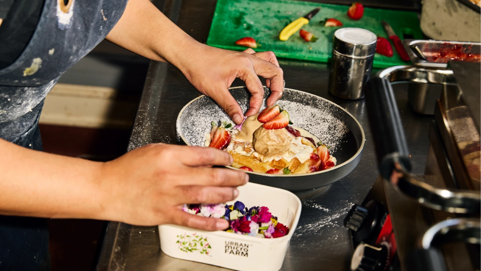 A chef carefully garnishes a dessert plate with sliced fresh strawberries and edible flowers in a commercial kitchen prep area.
