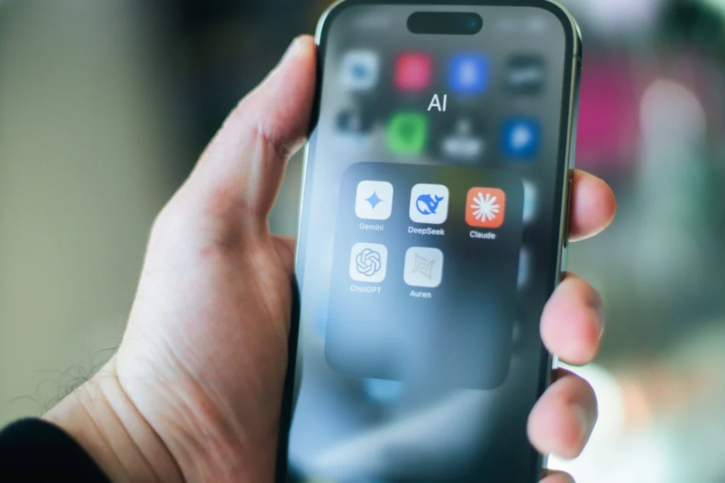 A hand holds a smartphone displaying an AI folder with apps like Gemini, DeepSeek, Claude, and ChatGPT.