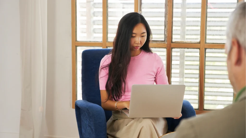 A young woman in a pink shirt sits in a blue armchair, focused on working on her laptop computer.