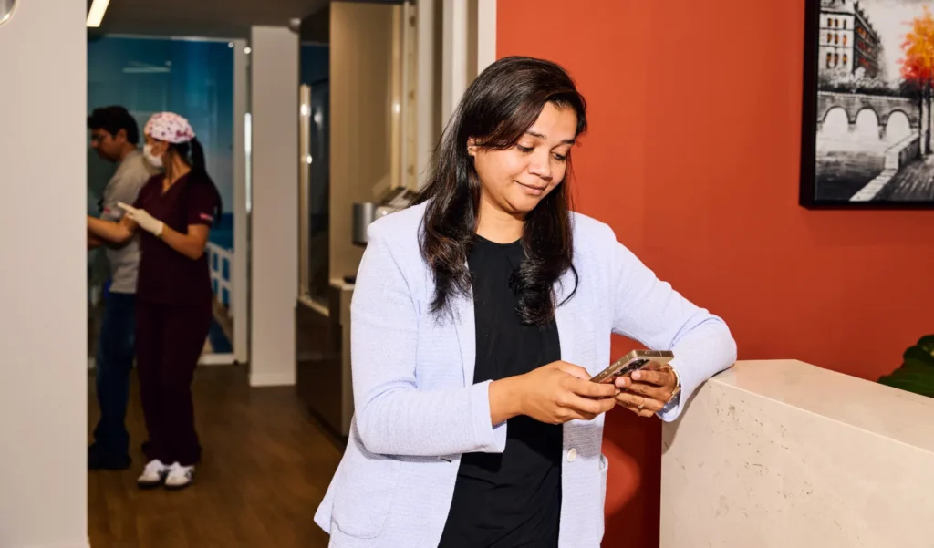 A woman in a light blue blazer using her smartphone while leaning against a counter in a medical clinic lobby.