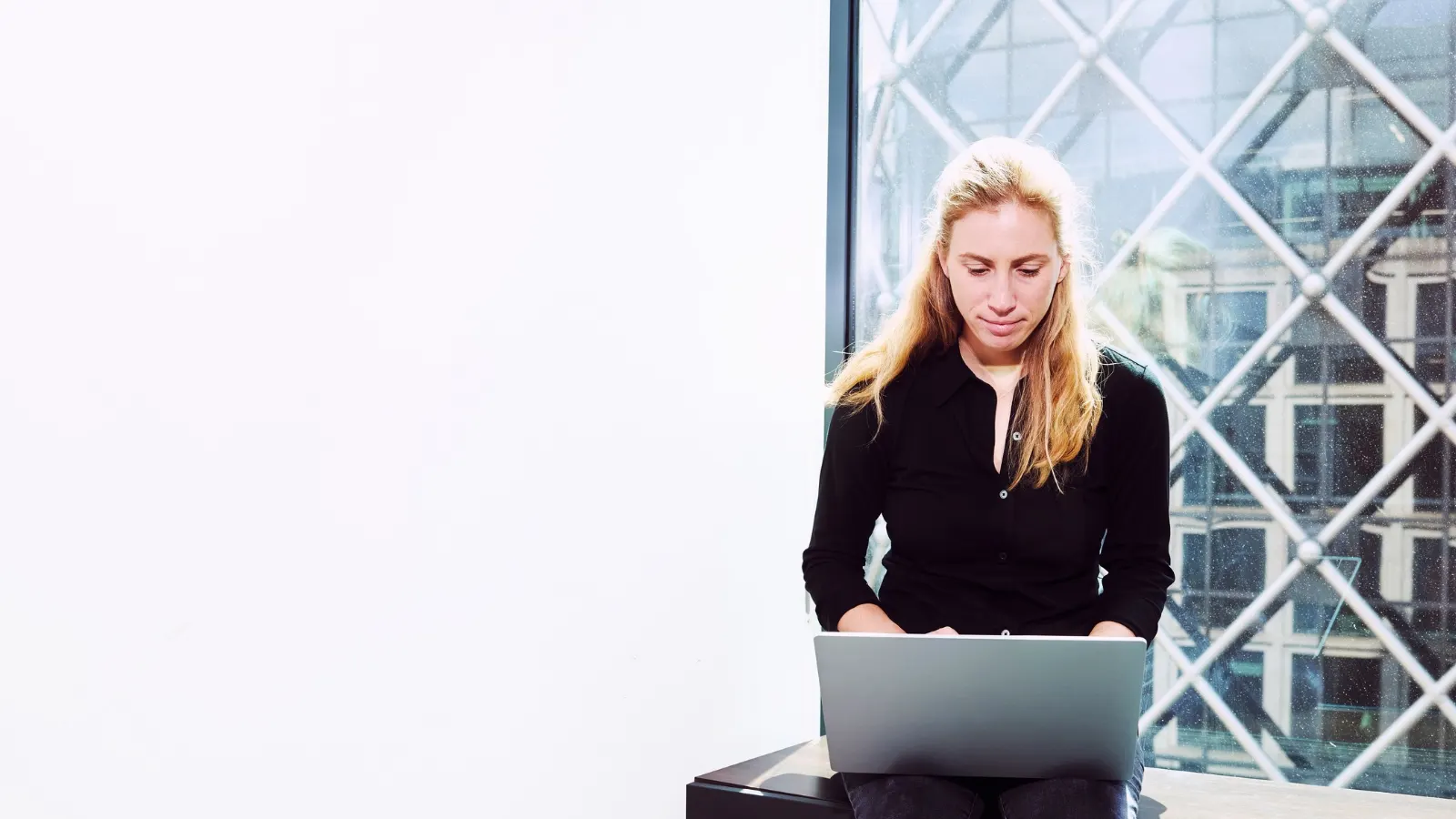 A blonde woman in a black shirt sits near a large glass window and types on a laptop.