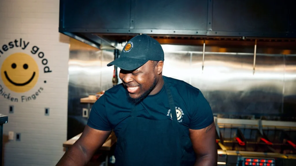 A smiling worker wearing a black t-shirt, apron, and cap featuring a smiley face logo stands in a commercial kitchen setting. Stainless steel kitchen equipment and branding with the phrase "honestly good" are visible around him.