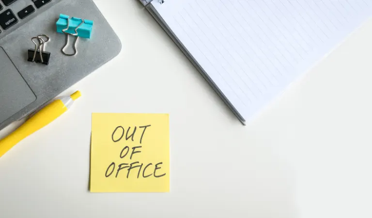 A top-down view of a white desk featuring a yellow sticky note that reads "OUT OF OFFICE" in handwritten capital letters.