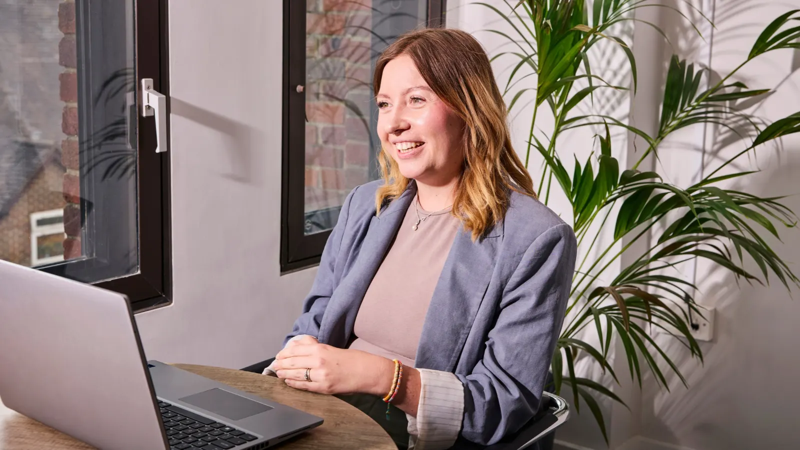A smiling woman in a blue blazer sits at a wooden desk with an open laptop next to a potted palm plant.