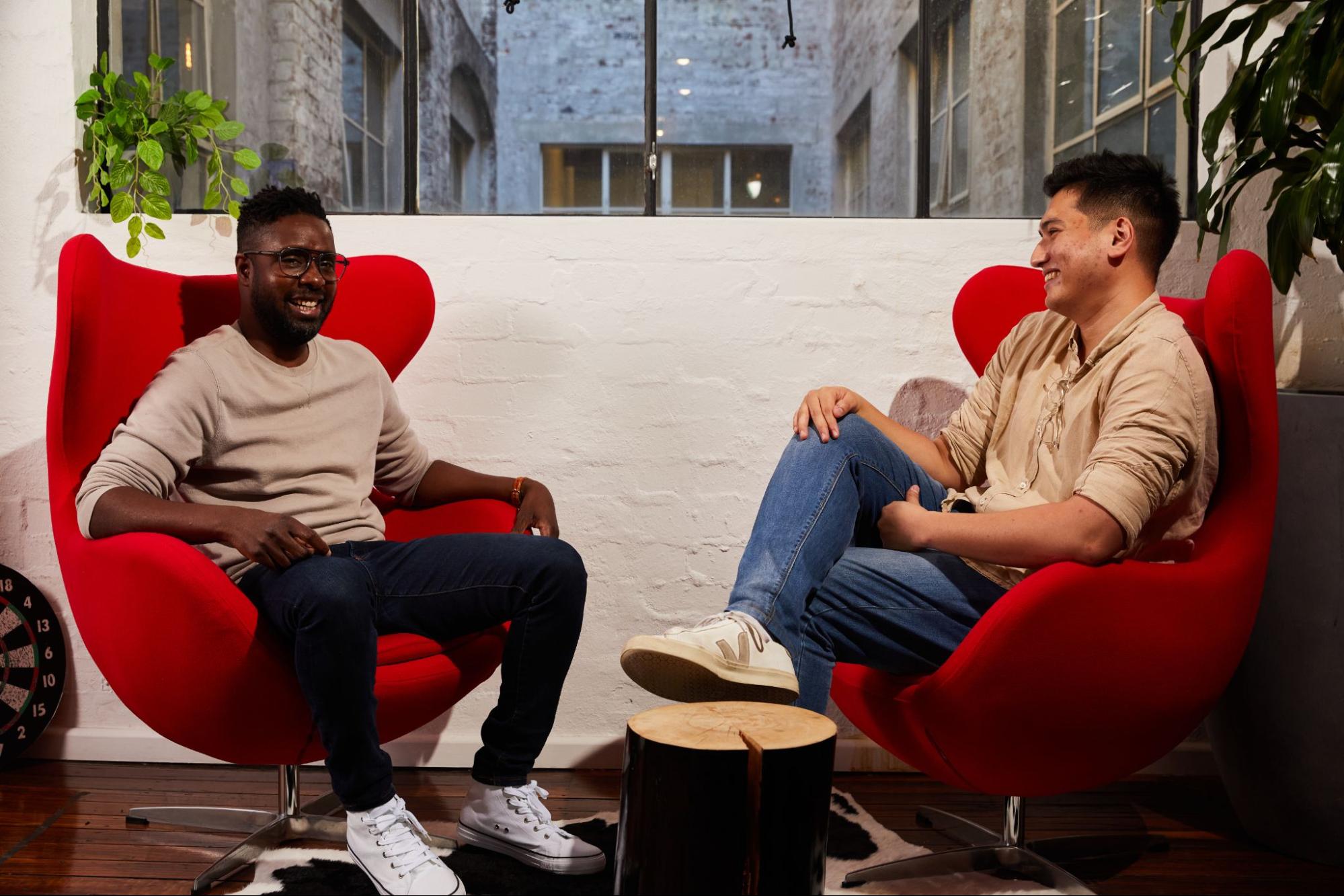 Two men are seated and conversing in red chairs within a modern room, featuring a white brick wall, plants, a wooden floor, and a dartboard.