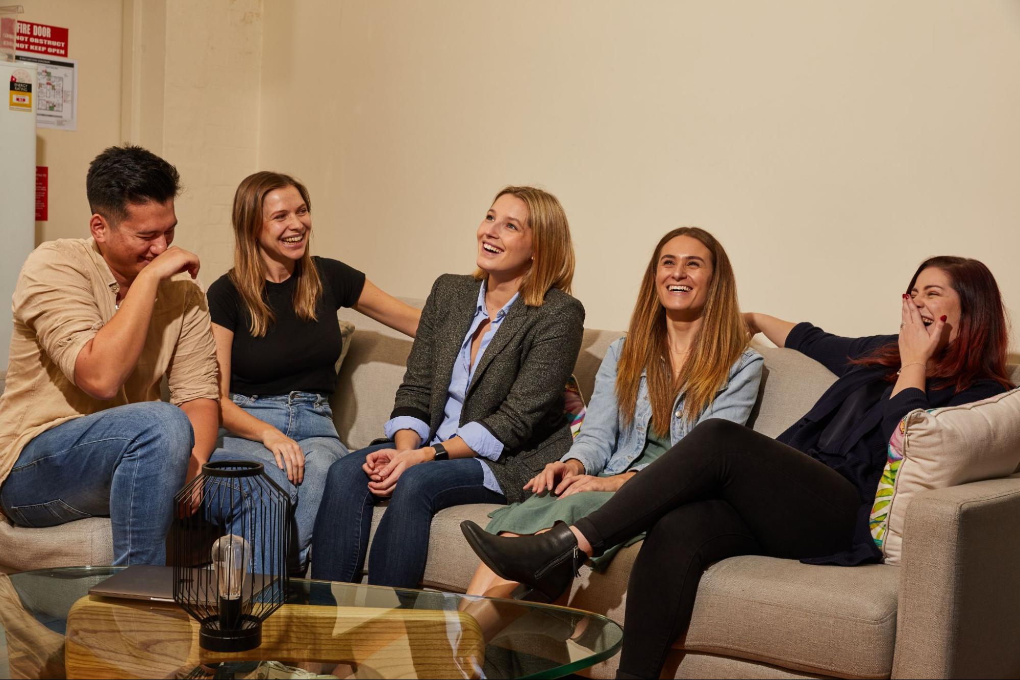 
Four people are laughing while sitting on a gray couch. The first person is a man with his right arm over his mouth, hiding his laughter, and his eyes closed. Next to him is a brunette woman wearing a loose black shirt and faded jeans. Beside her is a blonde woman with short hair, dressed in a blue shirt and a gray coat over dark blue skinny jeans. The fourth person is a woman wearing a light blue top over a green dress. She is sitting beside another woman, who has her hands on her lips, hiding her laughter, and is dressed entirely in black, from her shirt to her boots. In front of them is a glass table with an encased lightbulb as a centerpiece.
